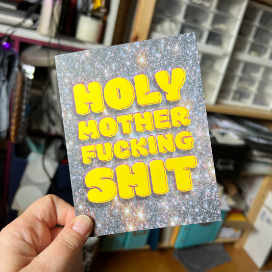 Hand holding a card with yellow text on a glittery background in a cluttered room.
