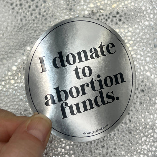 Metallic sticker with 'I donate to abortion funds' text held against a textured silver background