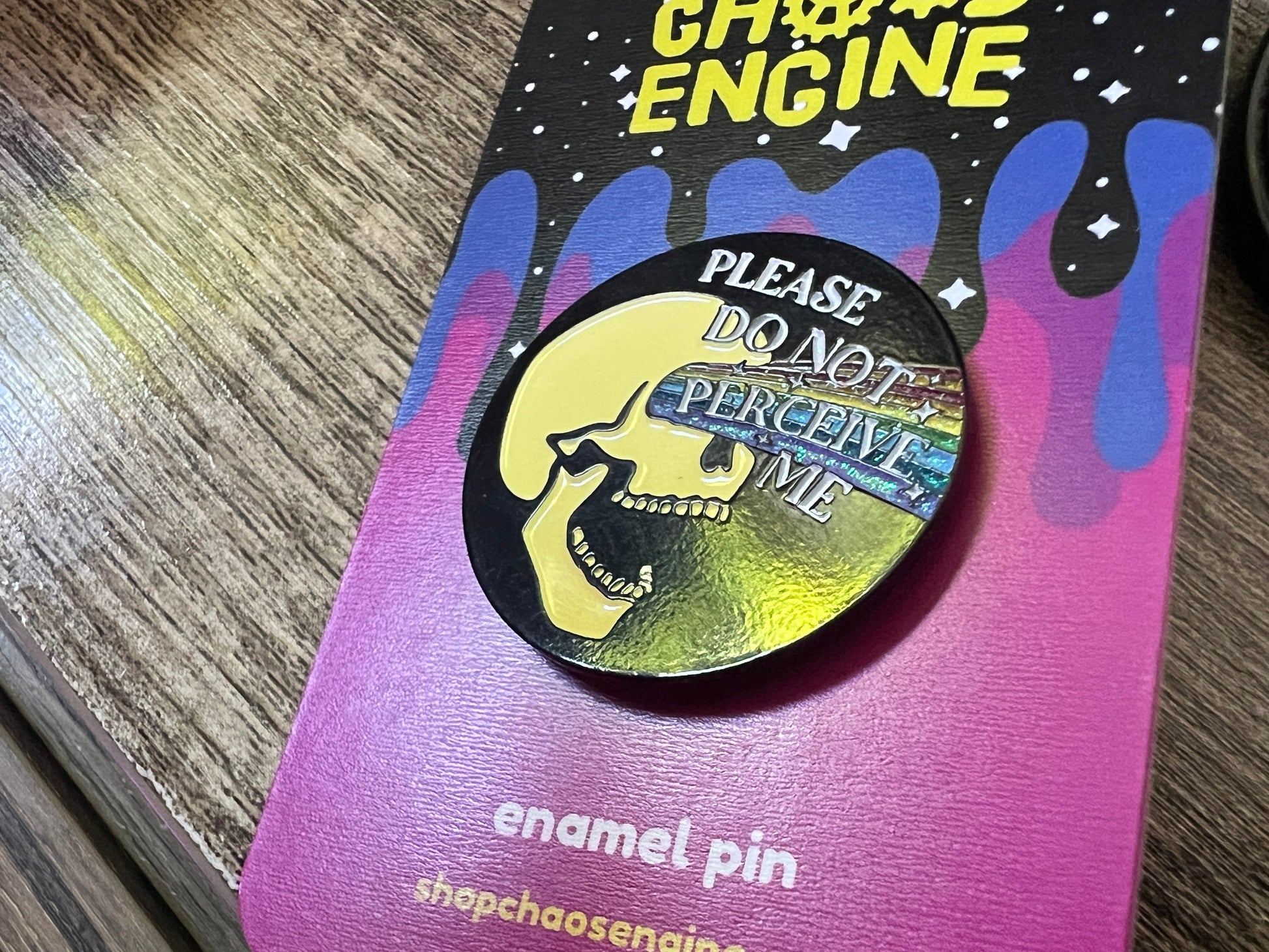 Enamel pin with skull design and text on a colorful card, brand 'Chaos Engine' visible.