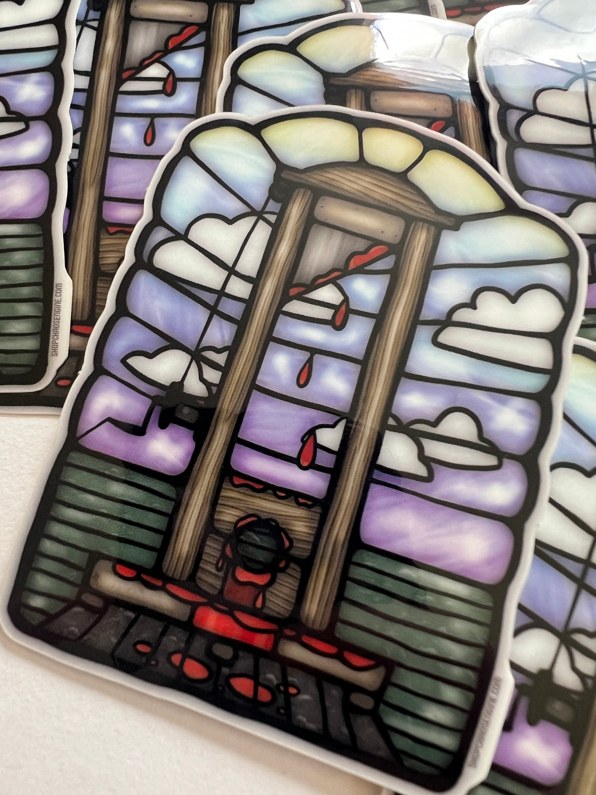 A stained-glass-style die-cut sticker featuring a wooden gallows with red dripping elements, white clouds, and purple sky panels, shown on a light surface with other similar stickers partially visible behind it.