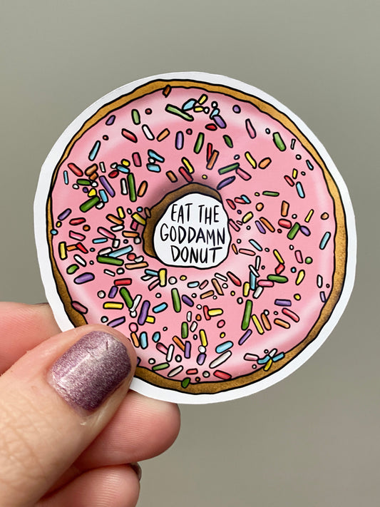 Sticker of a pink donut with sprinkles and 'Eat the Goddamn Donut' text held by a hand.
